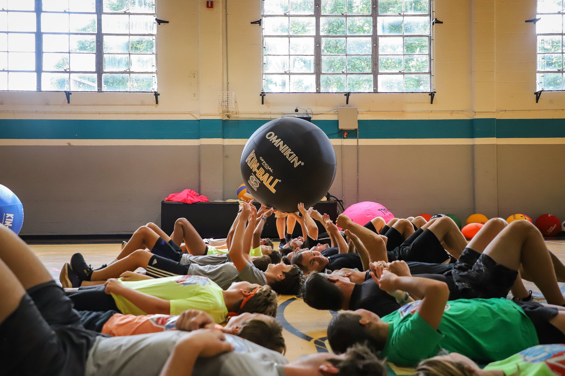 Students side by side laying on their backs passing an Omnikin Kin-Ball over their heads in a gymnasium.