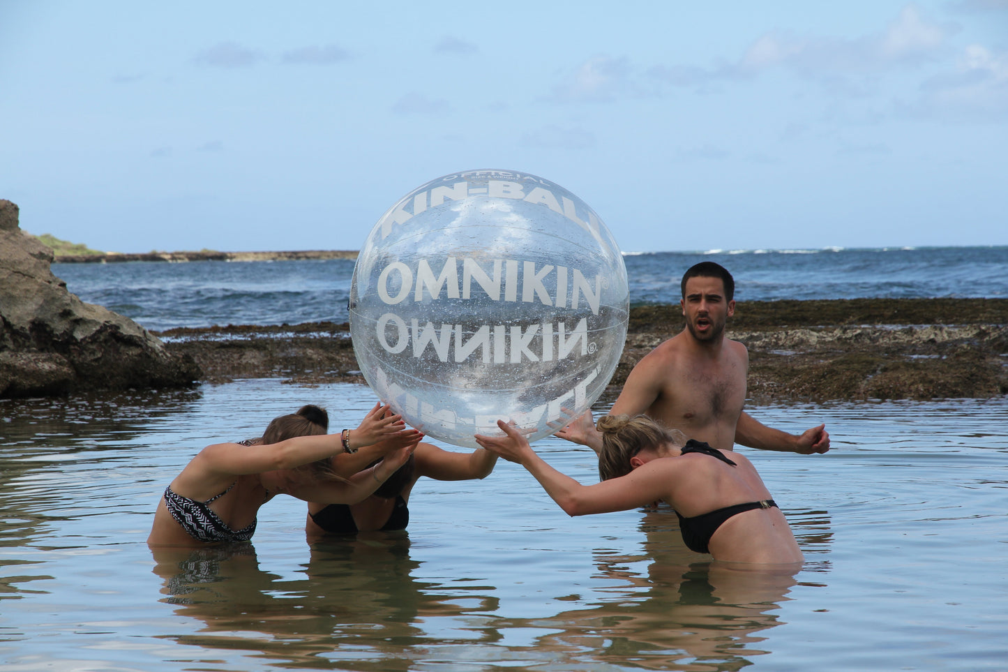 Three adults in a body of water holding a large, clear OMNIKIN ball above their heads. A Fourth adult is standing beside them to the right. The sky is blue with a few clouds.