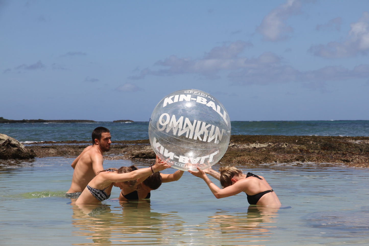 Three adults in a body of water holding a large, clear OMNIKIN ball above their heads. A Fourth adult is standing beside them. The sky is blue with a few clouds.