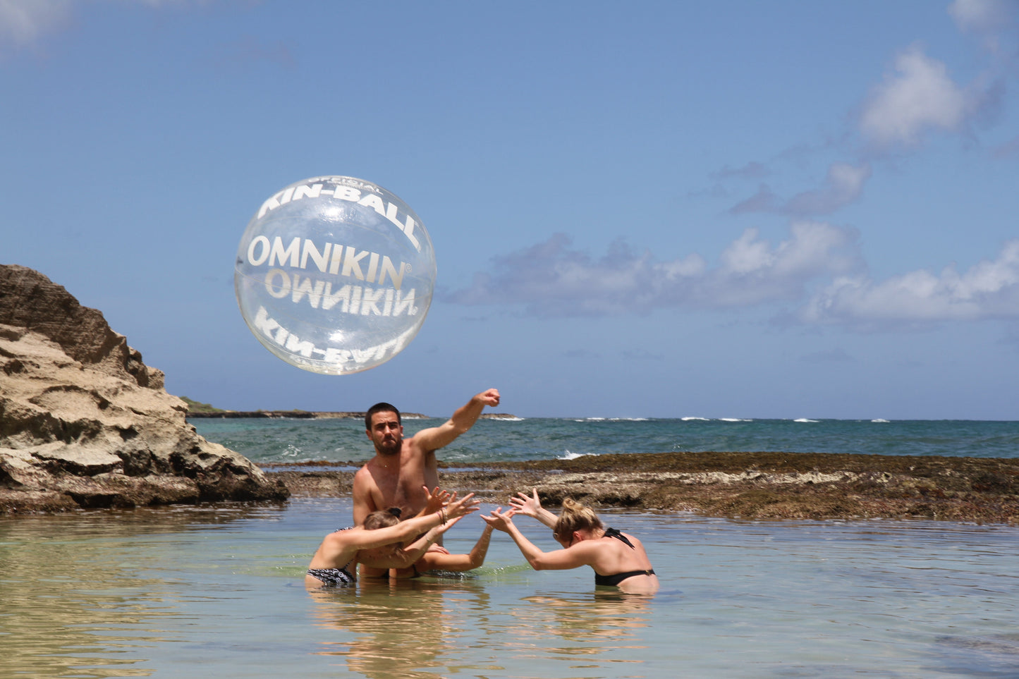 Three adults standing in a body of water. Two have their hands above their heads and the third is actively hitting a OMNIKIN KIN-BALL Street Sport Ball into the air.