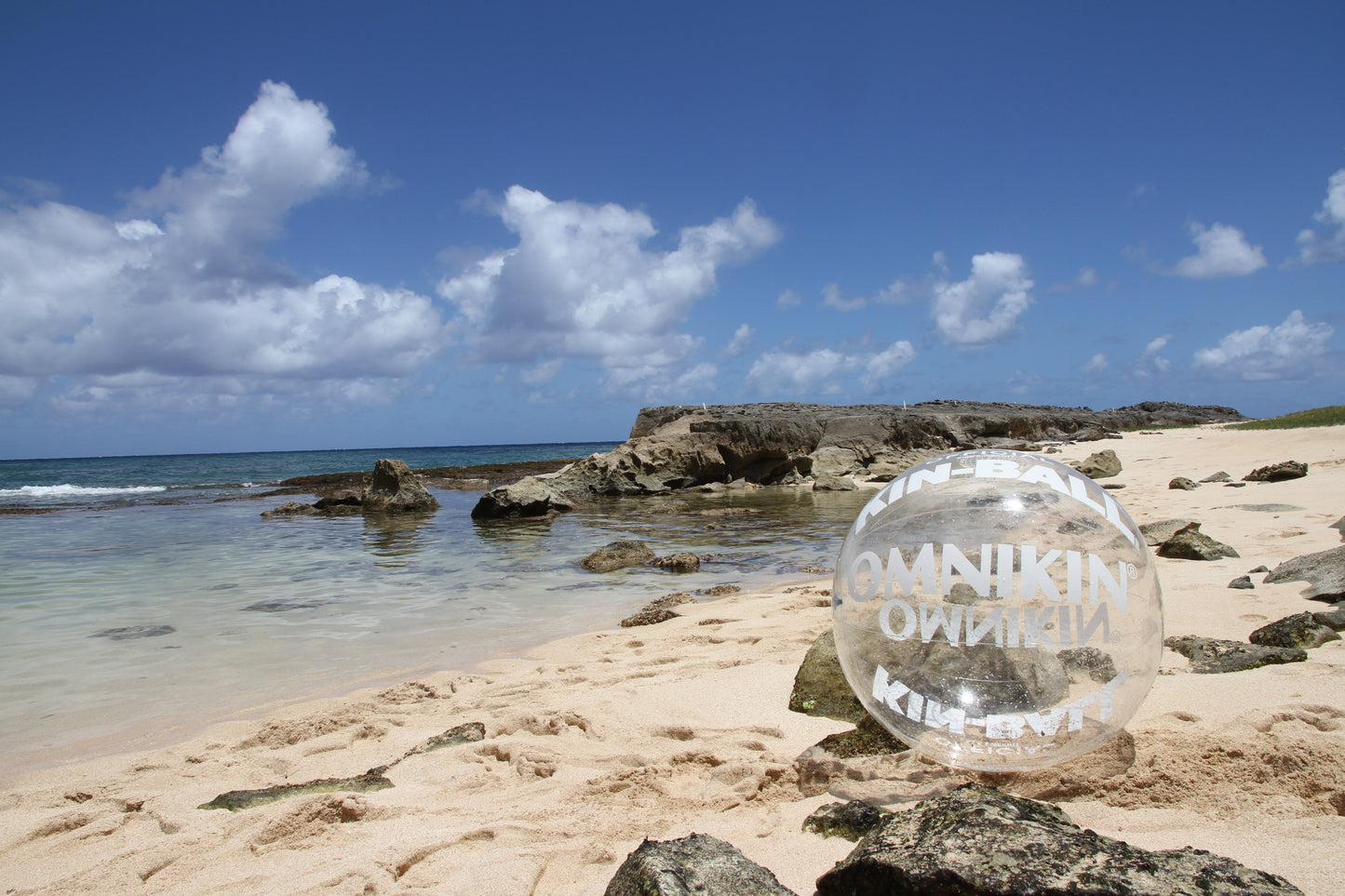 Clear OMNIKIN KIN-BALL Street Sport Ball on a beach with blue skies, clear water, sand and rocks.
