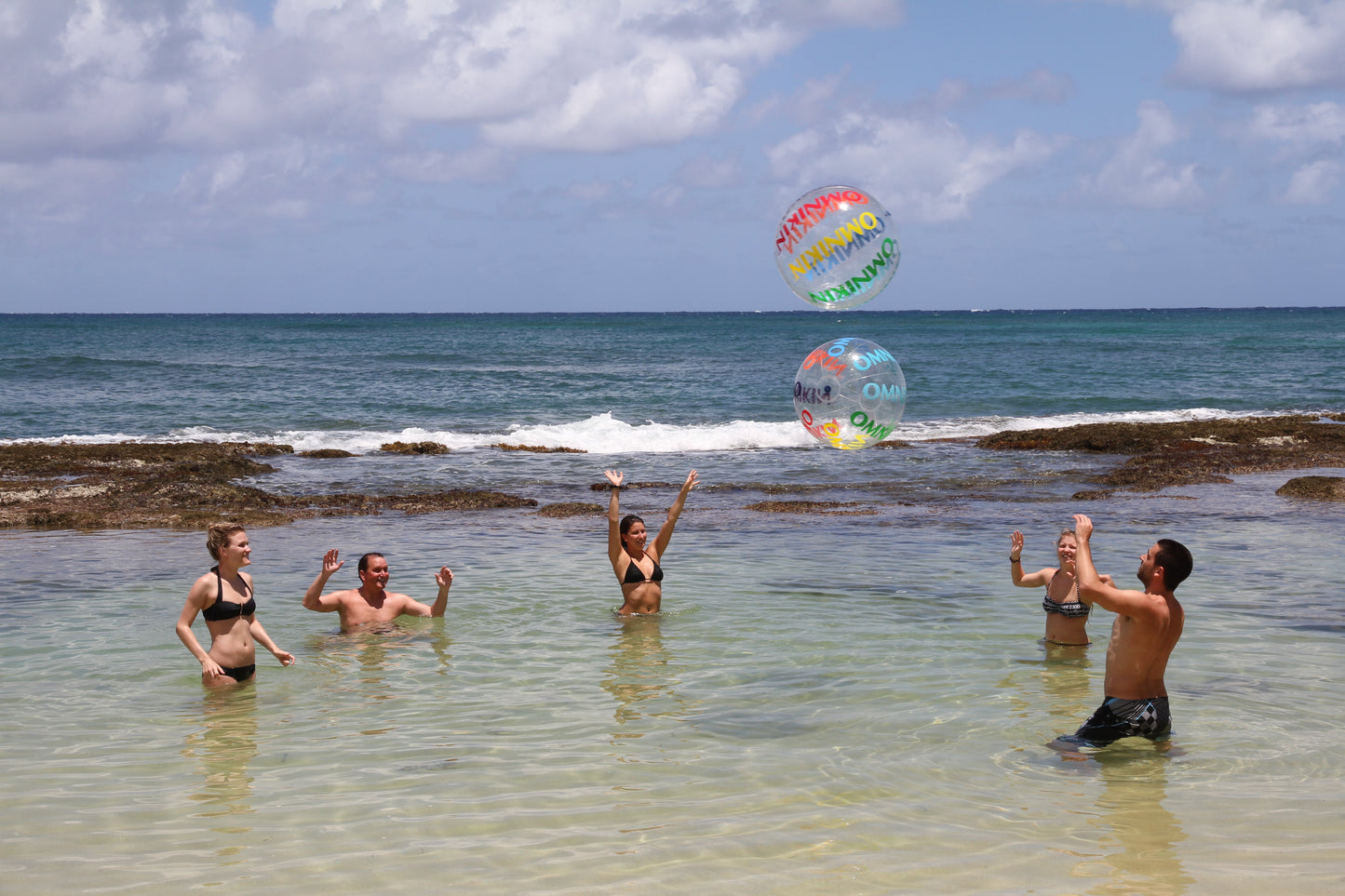 Adults in a body of water with blue skies overhead throwing two Omnikin Fit Balls up in the air.