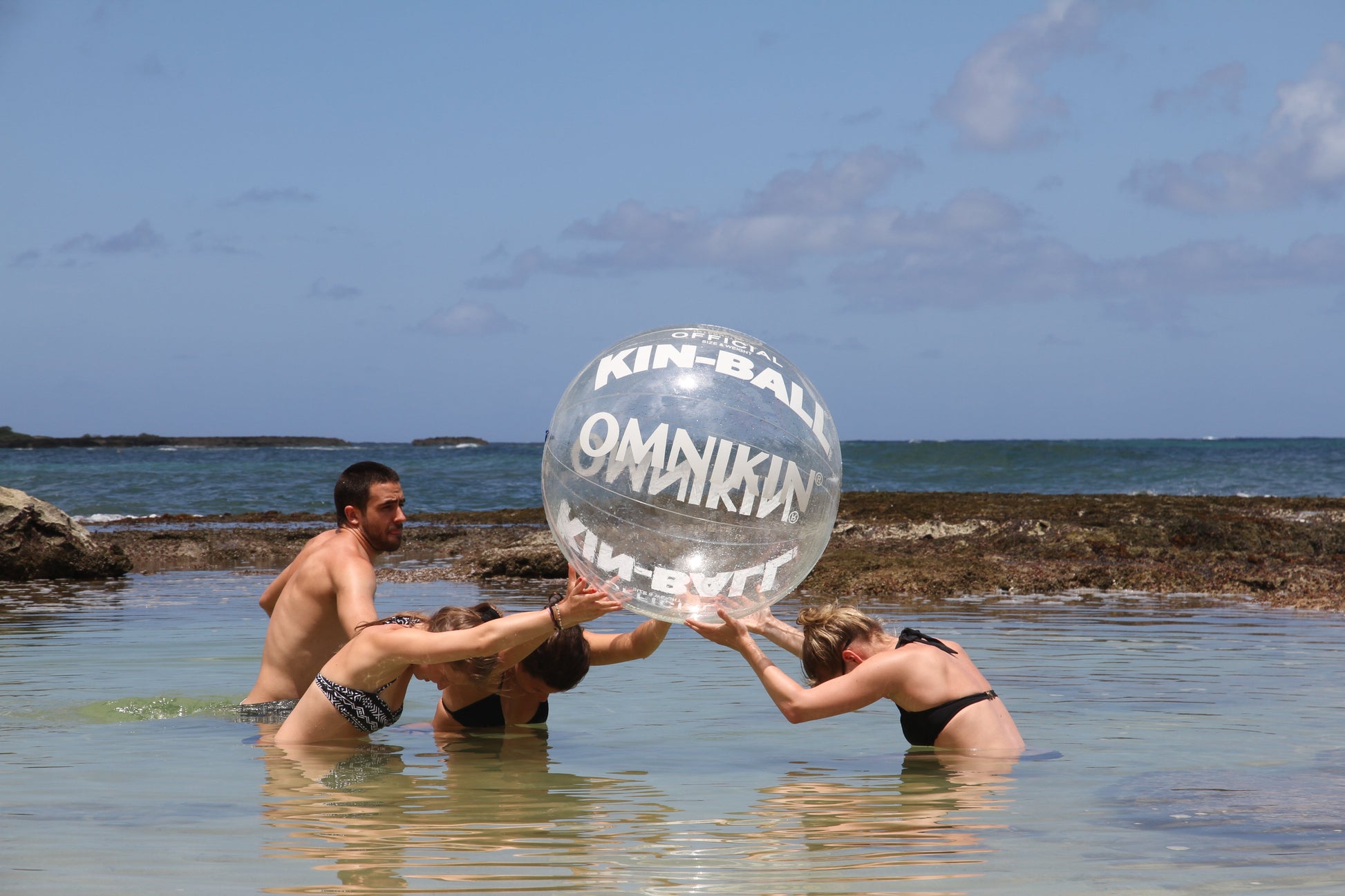 Three adults in a body of water holding a large, clear OMNIKIN ball above their heads. A Fourth adult is standing beside them. The sky is blue with a few clouds. 